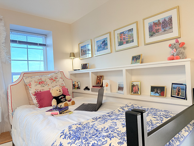 Stylish dorm bed with floral pillows and an emotional support teddy bear, laptop, and books, framed by a long headboard shelf displaying personal photos.