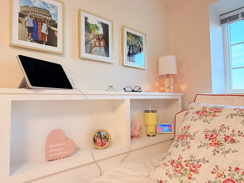 Cozy dorm bed setup featuring a floral pillow, framed photos, a tablet, and a yellow Yeti tumbler on a built-in white headboard shelf.