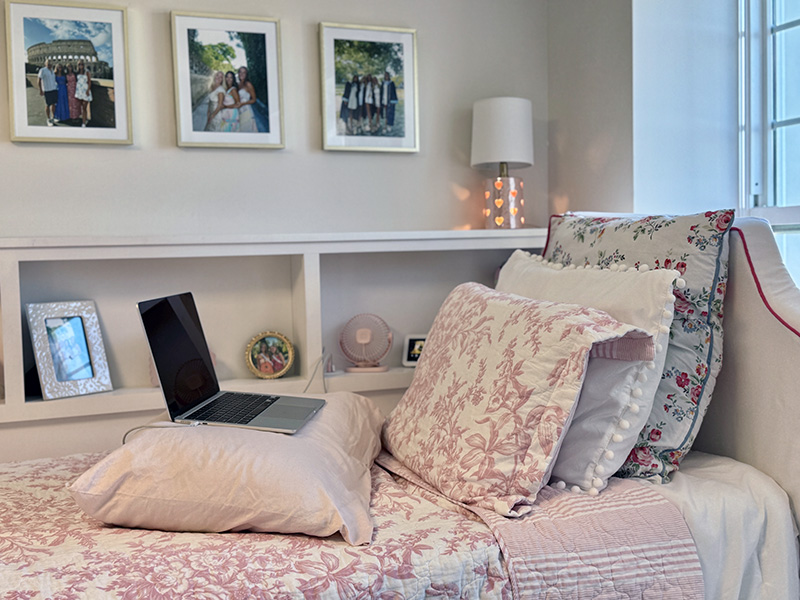 Laptop resting on a pillow atop a pink-accented dorm bed, with framed family photos and floral bedding against a cubby headboard.