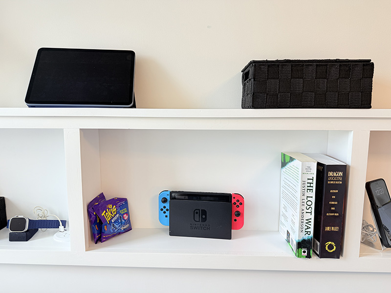Central cubby shelf filled with a Nintendo Switch, books, a snack pack, and a woven storage basket, with a tablet and decor above.